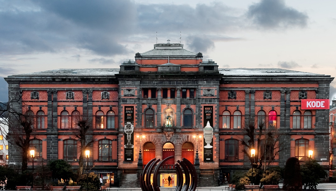 Frontal view of the facade of Permanenten (Kode) with street lights shinging in front and the cloudy sky above.