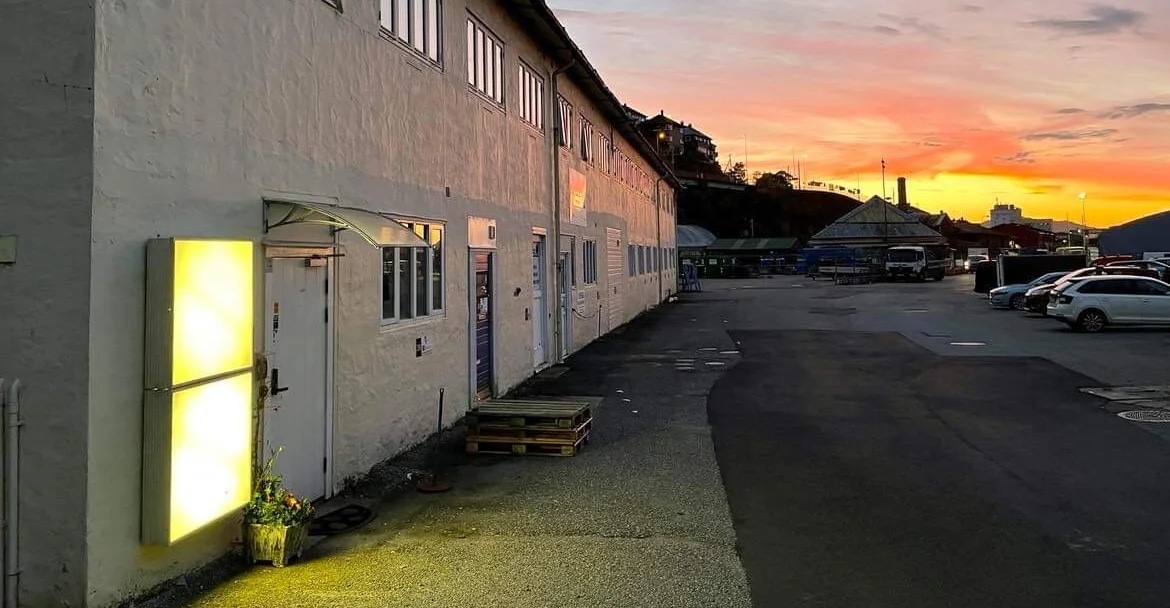 Picture of the entrance of the unfinished institute. An industrial building exterior with a glowing light panel at dusk. A few pallets are on the ground, and parked cars are visible in the background under a colorful sunset sky.