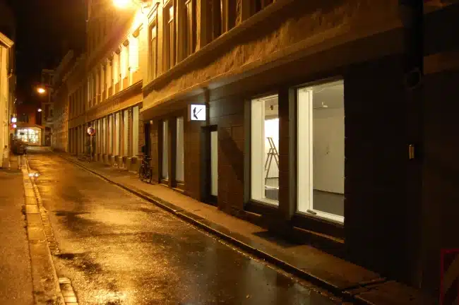 A dimly lit, wet and empty street at night. It is Østre Skost Brightly lit storefront windows line the right side of the street.