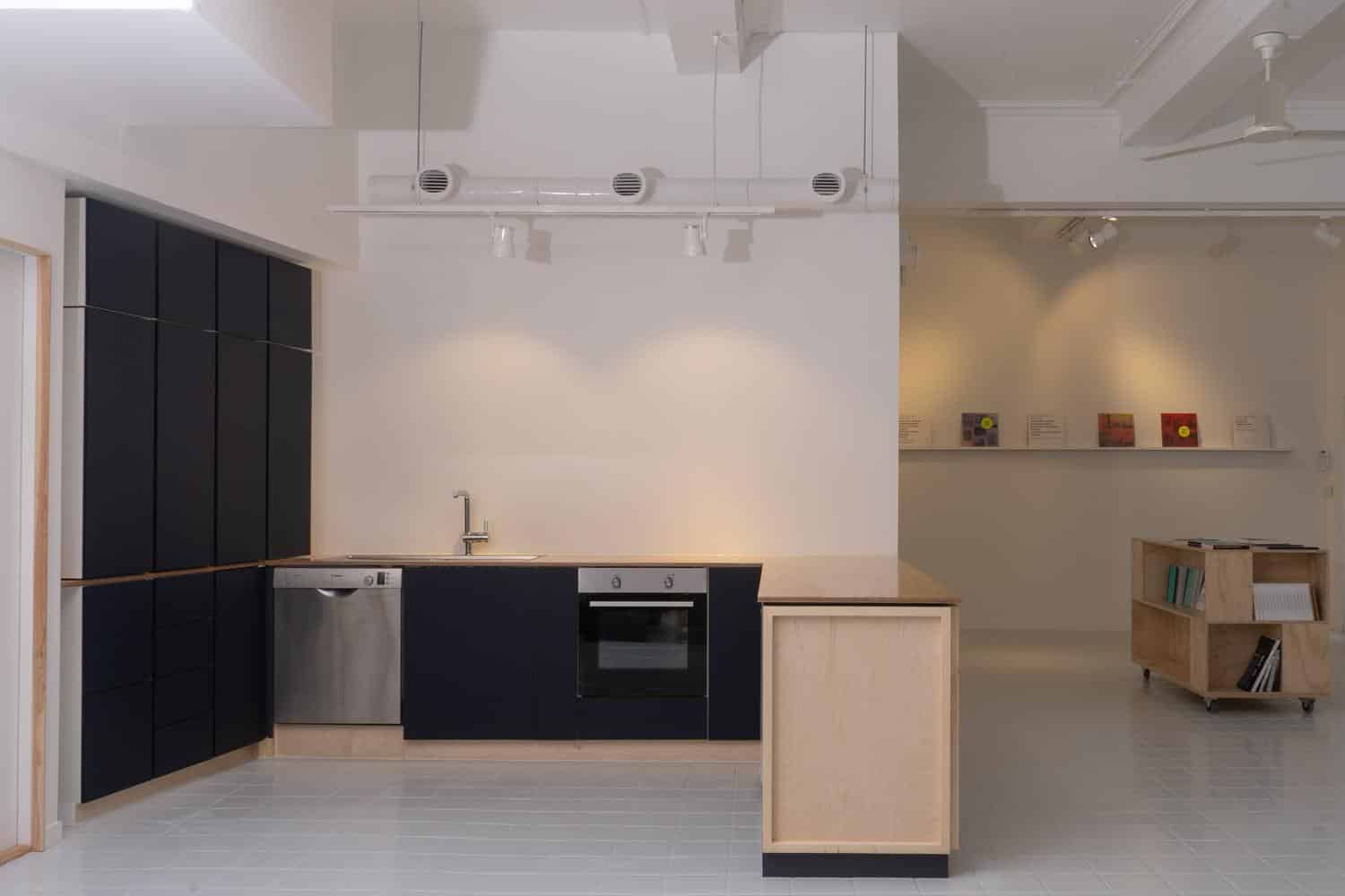 Minimalist kitchen of Bergen Assembly with black cabinets, stainless steel appliances, and a wooden countertop. Bright space with white walls and a small display area in the background.